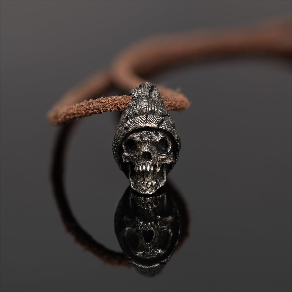 Beanie skull necklace standing on dark reflective glass with its mirror image, nickel silver hatted skull pendant on a leather cord