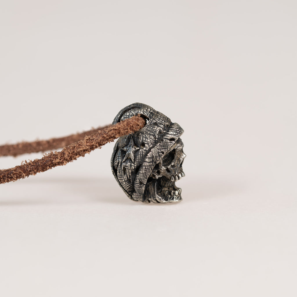 Side profile of the beanie skull pendant lying on a light surface, folded brim over the skull face, leather cord through the top of the hat