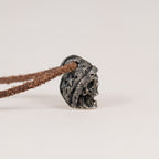 Side profile of the beanie skull pendant lying on a light surface, folded brim over the skull face, leather cord through the top of the hat