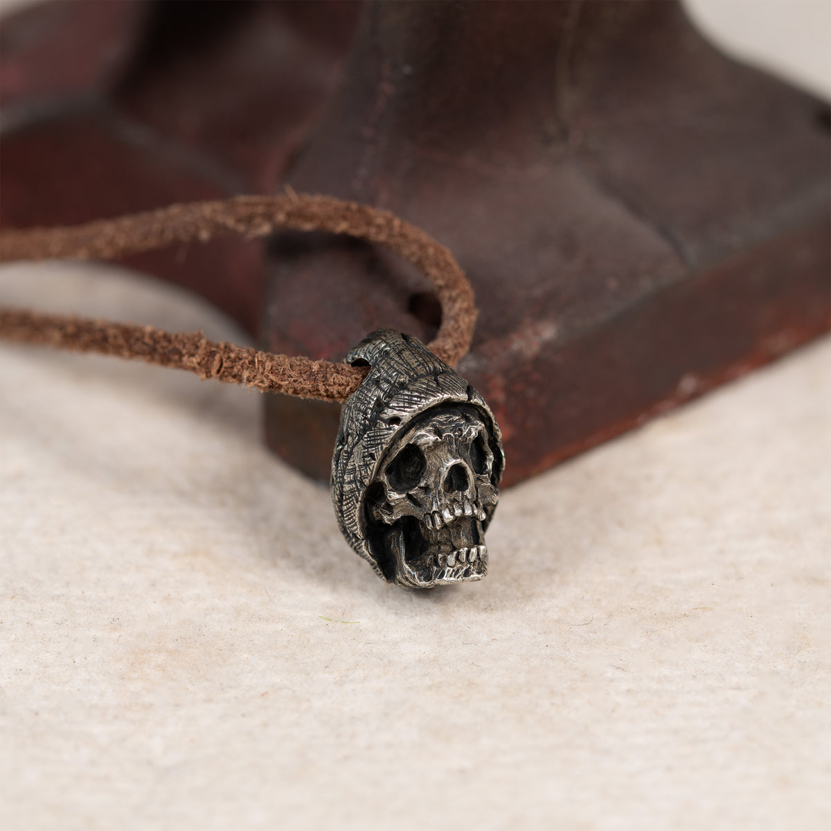 Beanie skull pendant resting against a rusted red iron tool on cream felt, leather cord, gothic memento mori necklace