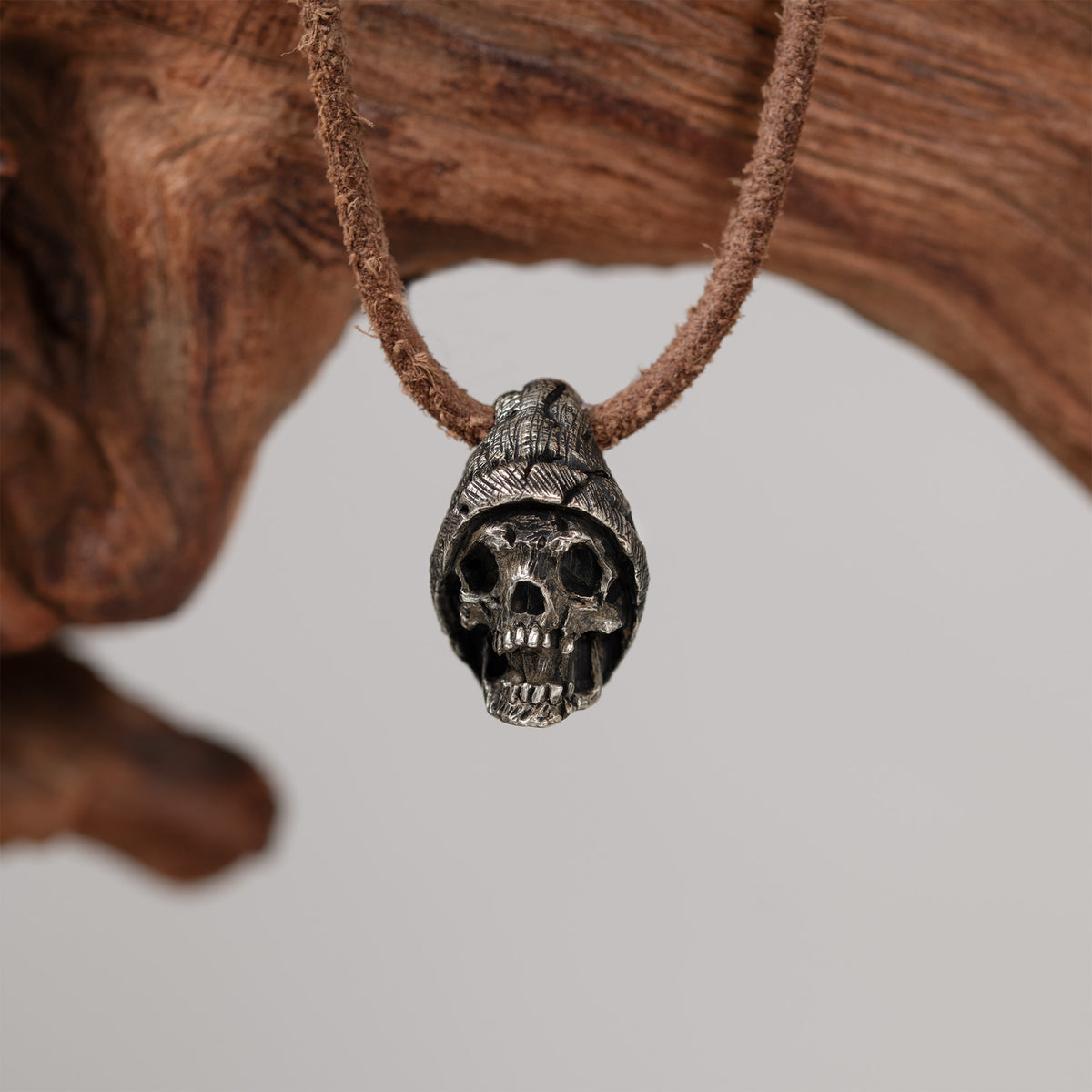 Beanie skull necklace hanging from a piece of driftwood, oxidized nickel silver pendant on a dark brown leather cord