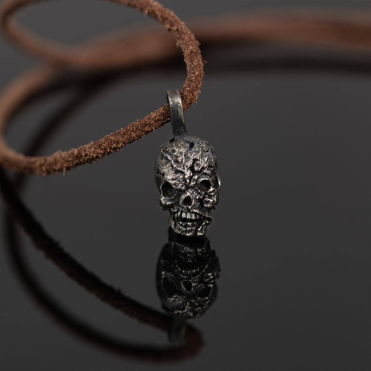 Exposed brain skull necklace standing on dark reflective glass with its mirror image, nickel silver skull pendant on a leather cord