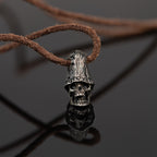 Top hat skull necklace standing on dark reflective glass with its mirror image, oxidized nickel silver skull pendant on a leather cord