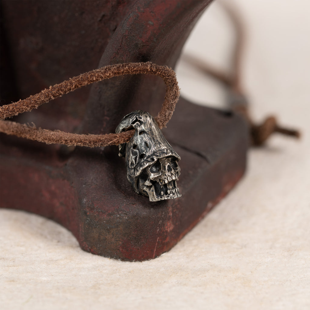 Top hat skull pendant resting against a rusted red iron tool on cream felt, leather cord, gothic memento mori necklace