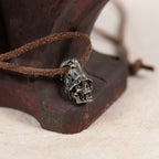 Top hat skull pendant resting against a rusted red iron tool on cream felt, leather cord, gothic memento mori necklace