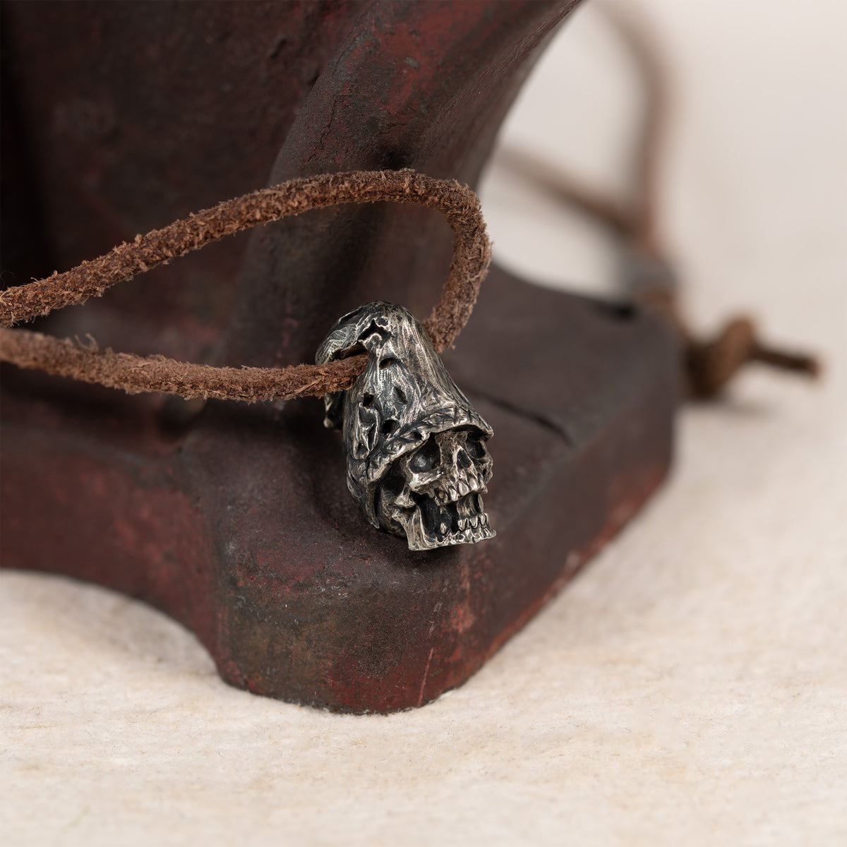 Top hat skull pendant resting against a rusted red iron tool on cream felt, leather cord, gothic memento mori necklace
