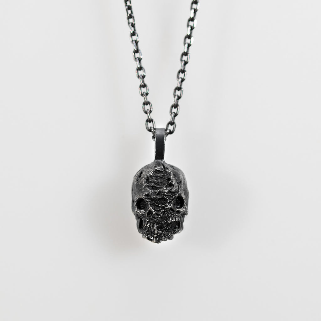 Double faced skull pendant in oxidized sterling silver hanging from a chain against a pale gray background.