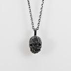 Double faced skull pendant in oxidized sterling silver hanging from a chain against a pale gray background.